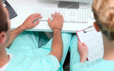 Two individuals in scrub suits seated at a desk, focused on a computer screen, engaged in a professional task.