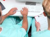 Two individuals in scrub suits seated at a desk, focused on a computer screen, engaged in a professional task.