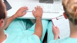 Two individuals in scrub suits seated at a desk, focused on a computer screen, engaged in a professional task.