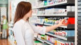  A woman examines a shelf filled with various medicine bottles in a well-lit pharmacy setting.