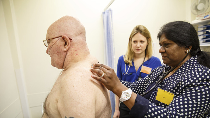 A group of healthcare professionals in a clinical setting, gathered around a table with laptops and medical documents. They are engaged in discussion, with one person pointing to a laptop screen and others looking on attentively. All are wearing professional attire, some with stethoscopes, indicating a medical or healthcare context