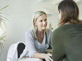 The image shows a healthcare professional, identifiable by the stethoscope around their neck and a white coat on the chair, consulting with a patient or client. They are seated across from each other at a table in what appears to be an office or clinical setting. The faces are obscured for privacy. This scene likely represents a medical consultation or therapy session.