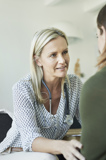 The image shows a healthcare professional, identifiable by the stethoscope around their neck and a white coat on the chair, consulting with a patient or client. They are seated across from each other at a table in what appears to be an office or clinical setting. The faces are obscured for privacy. This scene likely represents a medical consultation or therapy session.