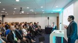  A man stands confidently at a podium, presenting to an engaged audience in a spacious conference hall.