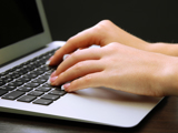 Close-up of hands typing on a laptop keyboard with a dark background. The person appears to have well-manicured nails, and the laptop is silver.