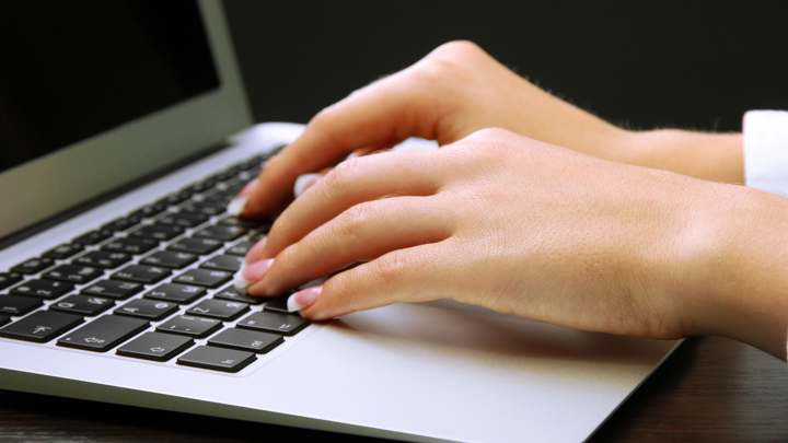 Close-up of hands typing on a laptop keyboard with a dark background. The person appears to have well-manicured nails, and the laptop is silver.