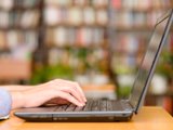 Individual engaged in typing on a laptop, positioned in front of a bookcase brimming with various books.